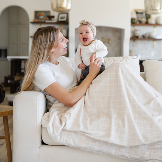 A mother holds her happy baby girl and is sitting with a luxuriously soft Saranoni minky/lush blanket. This premium dual-textured extra-large throw features Saranoni’s signature cream lush fabric on one side and ultra-soft square printed minky on the other. Designed for ultimate relaxation, this plush blanket is perfect for snuggling, movie nights, and adding a touch of luxury to any home. This soft blanket is a luxury blanket and a Saranoni blanket. The cozy blanket is a comfortable blanket.