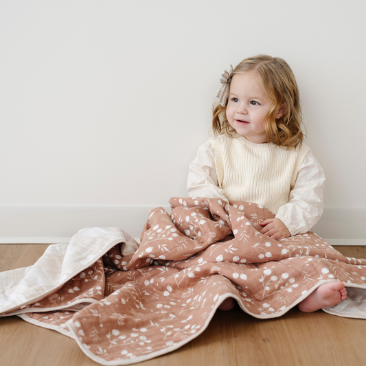 A little girl sits with a cotton muslin quilt that is a light brown with a floral pattern. This soft blanket is a great baby blanket or toddler blanket. It's adorable pattern makes it a great addition to a nursery or bedroom. Enjoy this Saranoni blanket for years to come.