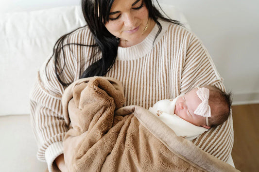 A mother in a warm neutral striped sweater holds her newborn wrapped in a soft taupe blanket, smiling down at her baby in a bright, minimalist room.