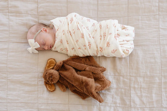 Newborn swaddled in a Saranoni blanket lies peacefully next to a soft brown stuffed bunny on a neutral quilted surface, capturing a moment of newborn comfort and gentle companionship.