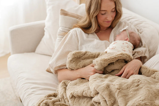 A mother sits on a cream bed holding her newborn, wrapped in a Saranoni Lush blanket, surrounded by soft, neutral textures and natural light.