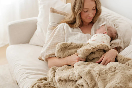 A mother sits on a cream bed holding her newborn, wrapped in a Saranoni Lush blanket, surrounded by soft, neutral textures and natural light.