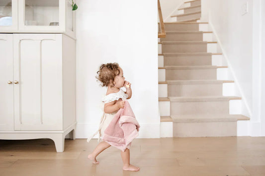 A toddler with curly hair runs across a light wood floor in a bright, minimalist home, wrapped in a pink Saranoni blanket.
