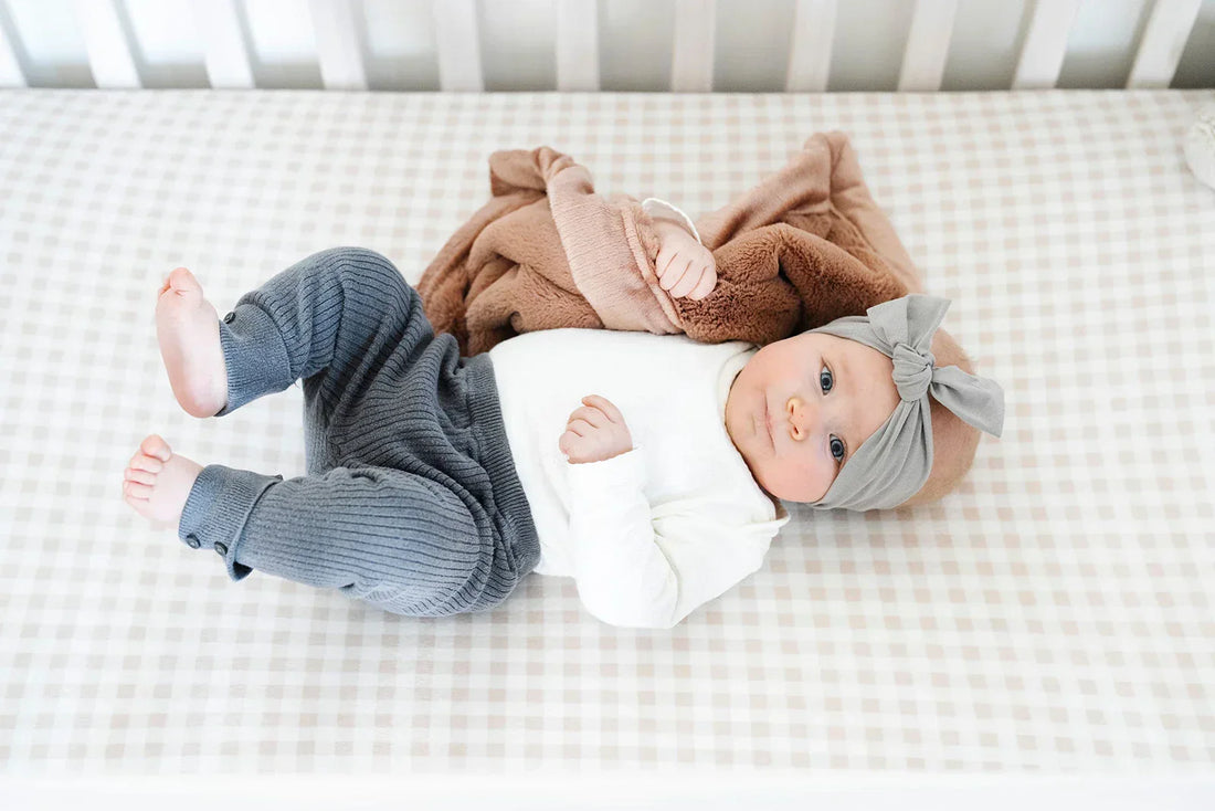 A baby lies on a white gingham crib sheet, wrapped in a warm brown Saranoni blanket, with a stuffed animal nearby in a bright, softly lit nursery.