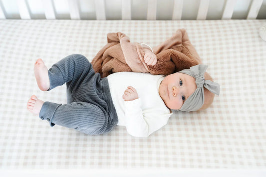 A baby lies on a white gingham crib sheet, wrapped in a warm brown Saranoni blanket, with a stuffed animal nearby in a bright, softly lit nursery.