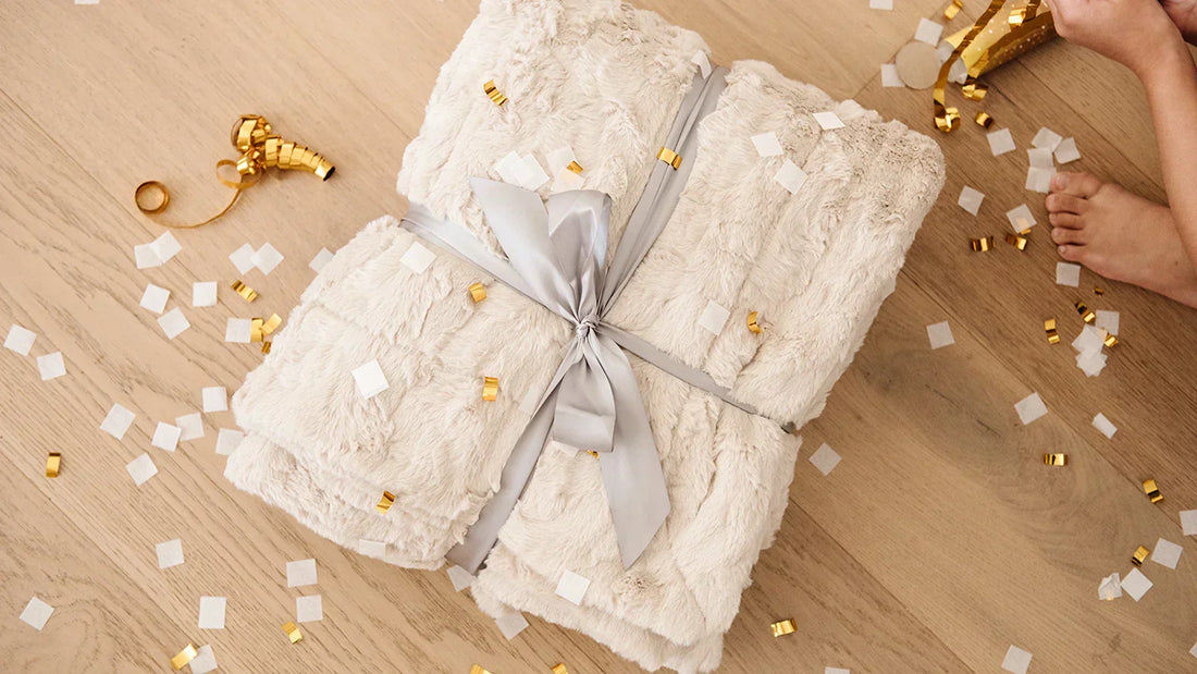 Ivory Lush blankets tied with a gray ribbon, surrounded by scattered gold and white confetti on a natural wood surface, with a hand reaching in from the right.