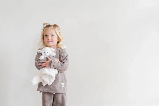 Toddler in a gray ribbed outfit holding a white Saranoni lovey against a plain wall