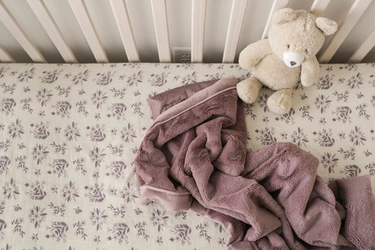 Mauve Saranoni blanket with satin border on a crib sheet with a lilac floral print beside a cream teddy bear