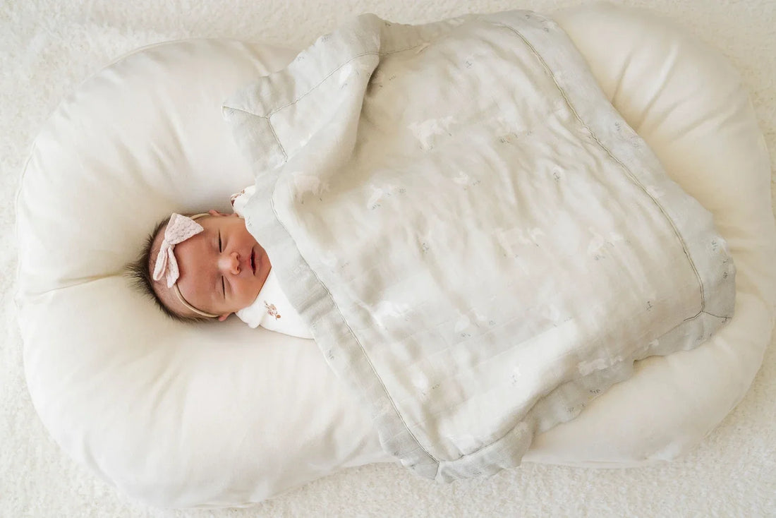 A newborn baby sleeps peacefully on a soft cushioned lounger, wrapped in a Saranoni blanket in gentle neutral tones.