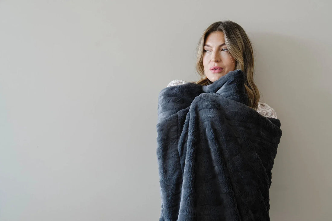 Woman in a cream knit sweater sits on a light gray window seat wrapped in a caramel chunky knit blanket, gazing thoughtfully toward natural light streaming through large windows.