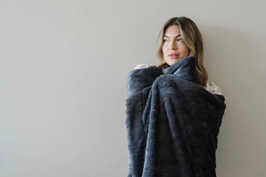Woman in a cream knit sweater sits on a light gray window seat wrapped in a caramel chunky knit blanket, gazing thoughtfully toward natural light streaming through large windows.