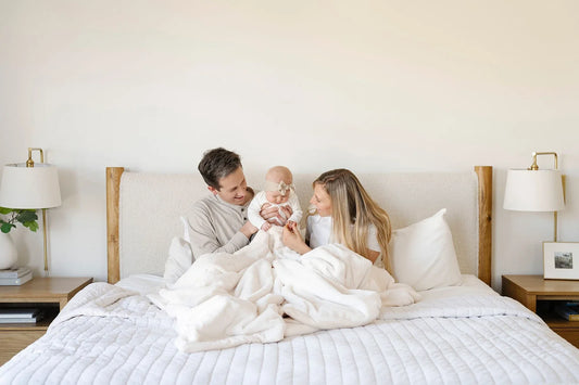 A family of four sits together on a bed wrapped in white Saranoni blankets, sharing a warm moment of connection and comfort.