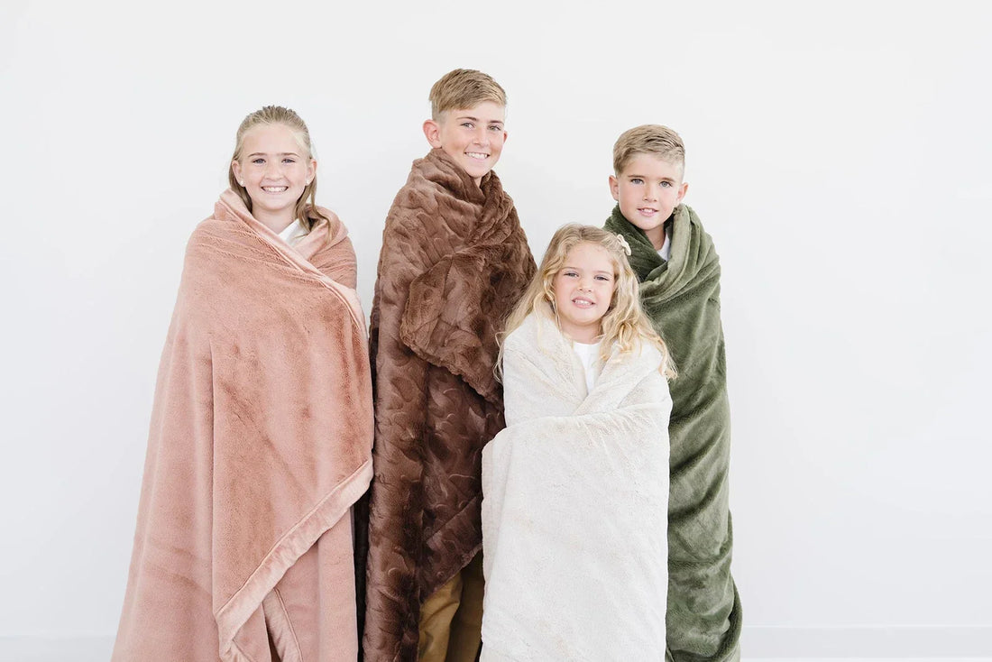 Four children wrapped in soft Saranoni blankets in warm neutral tones, smiling against a white background — a moment of family belonging.