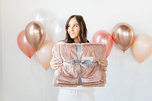 Woman holding a folded blush pink Saranoni blanket tied with a gray ribbon, surrounded by rose gold and blush balloons
