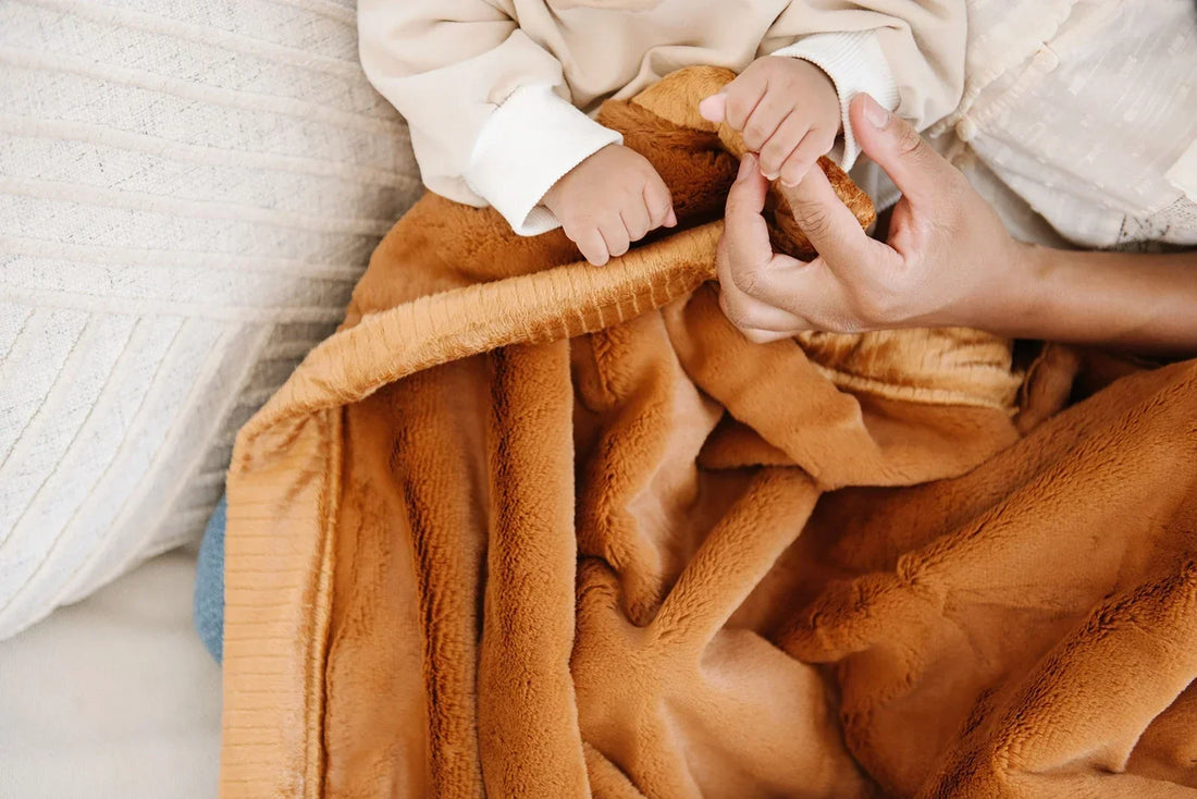 A baby's hands grip a caramel-toned Saranoni blanket while a parent holds the child close in a warm, intimate moment.