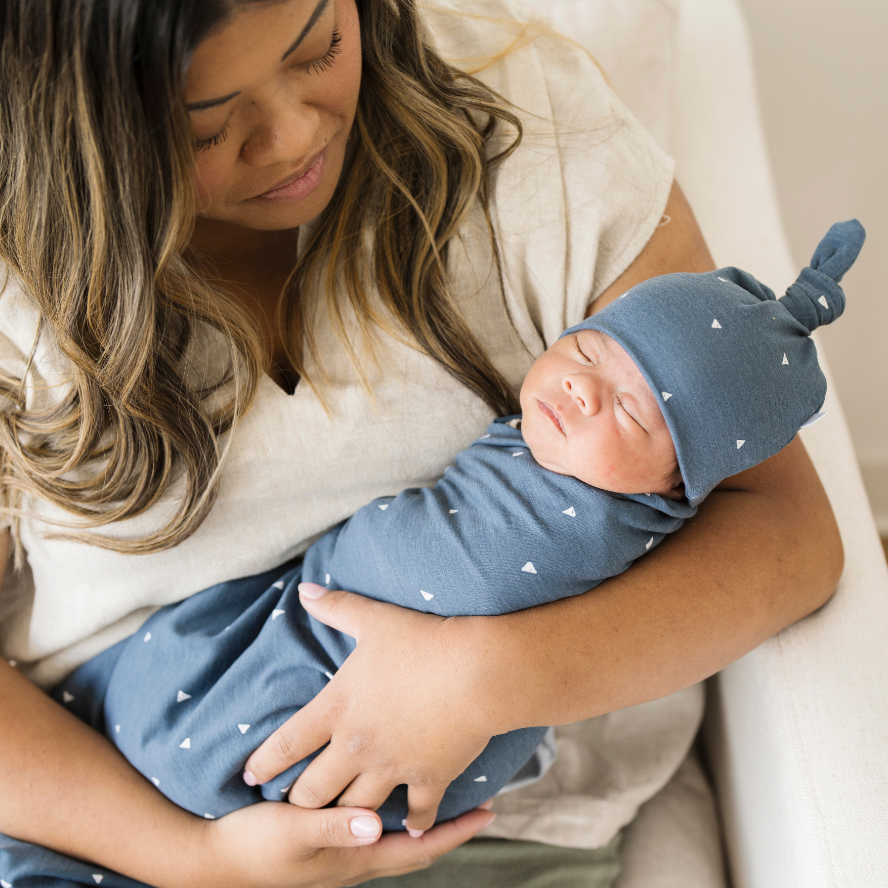 Mom holding baby boy swaddled in Saranoni Stretchy Swaddle in Denim Triangle with a matching hat. 