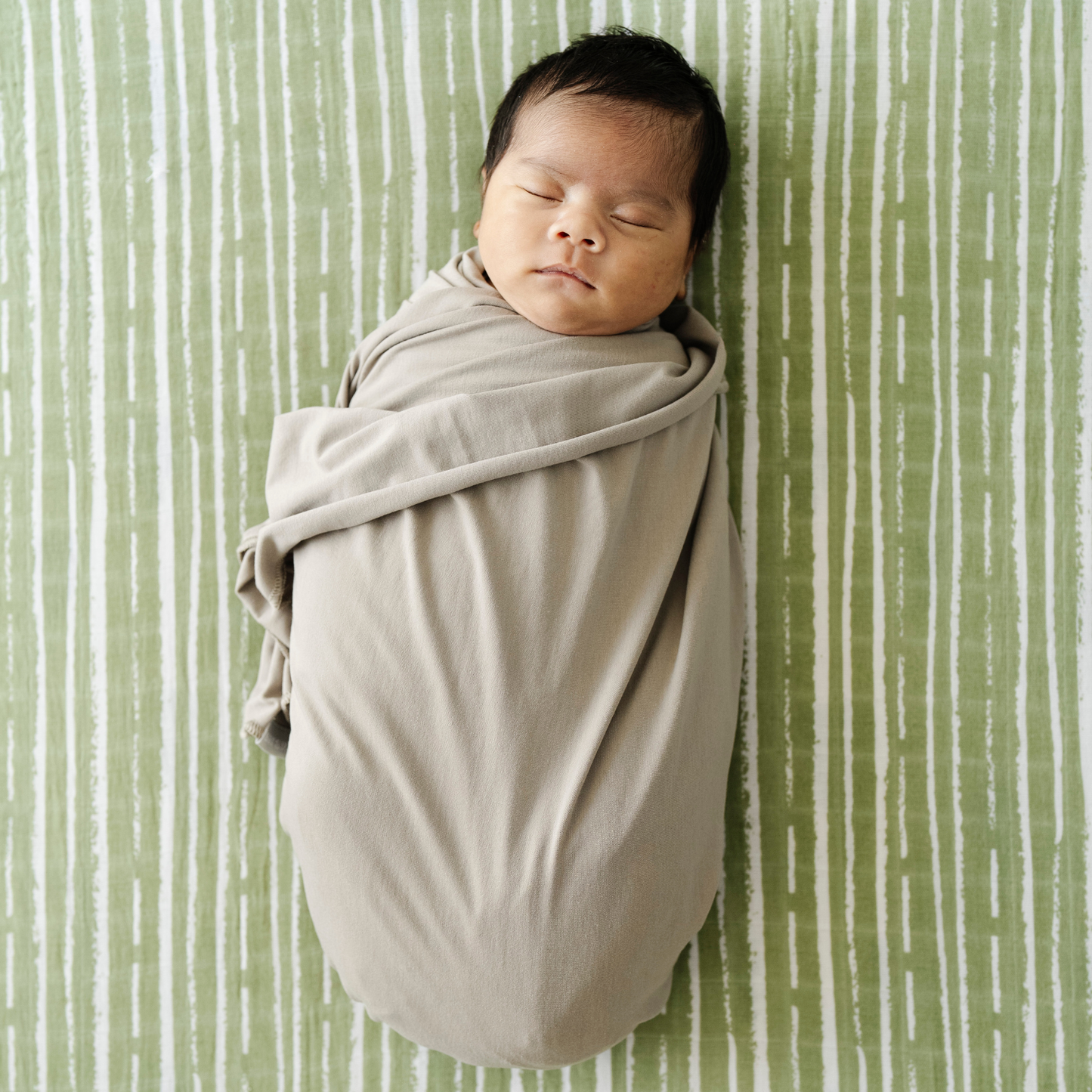Baby boy swaddled in Saranoni Stretchy Swaddle while laying on Forest Stripe Cotton Muslin Crib Sheet.
