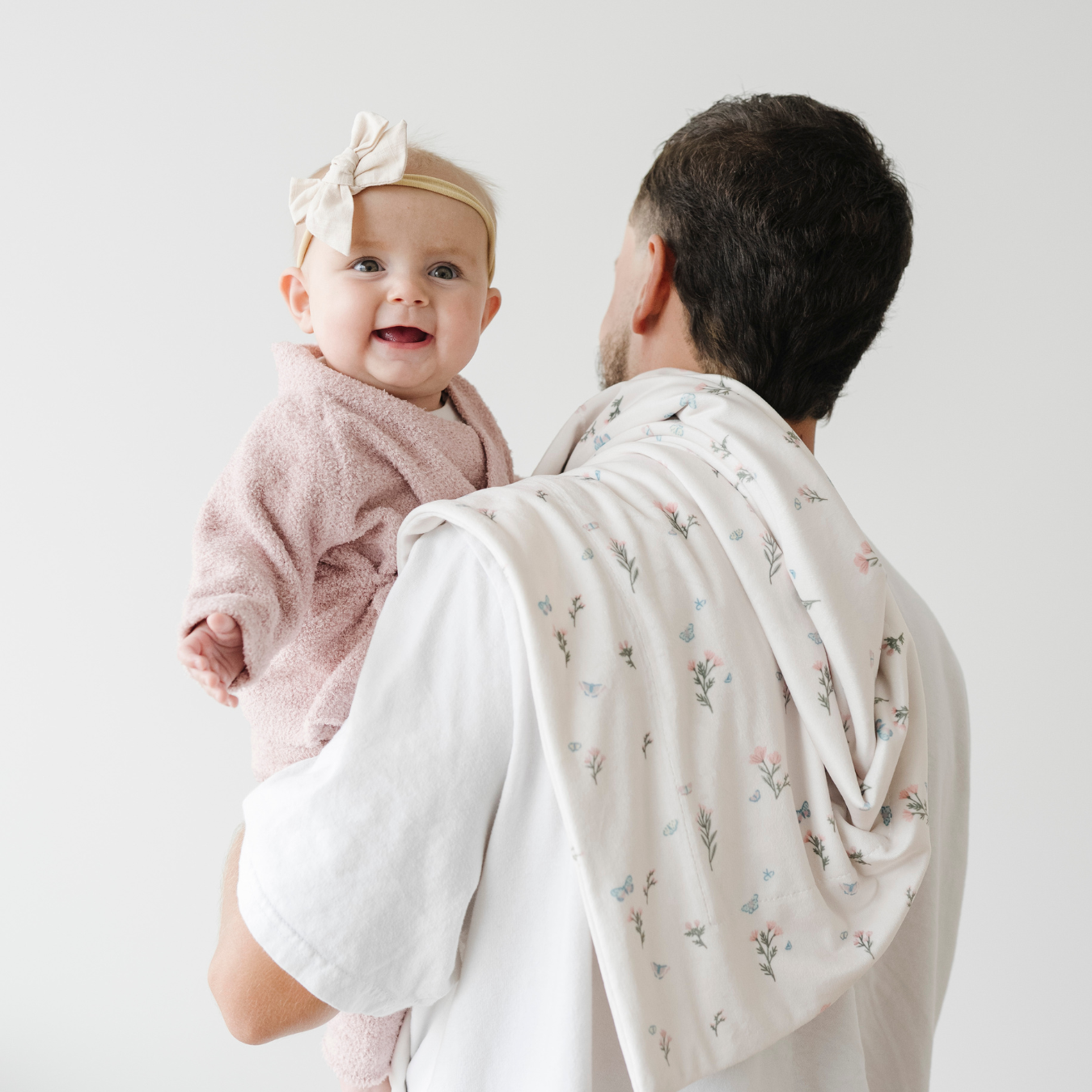 Baby in a pink outfit with a bow, held by dad holding a floral-patterned blanket.