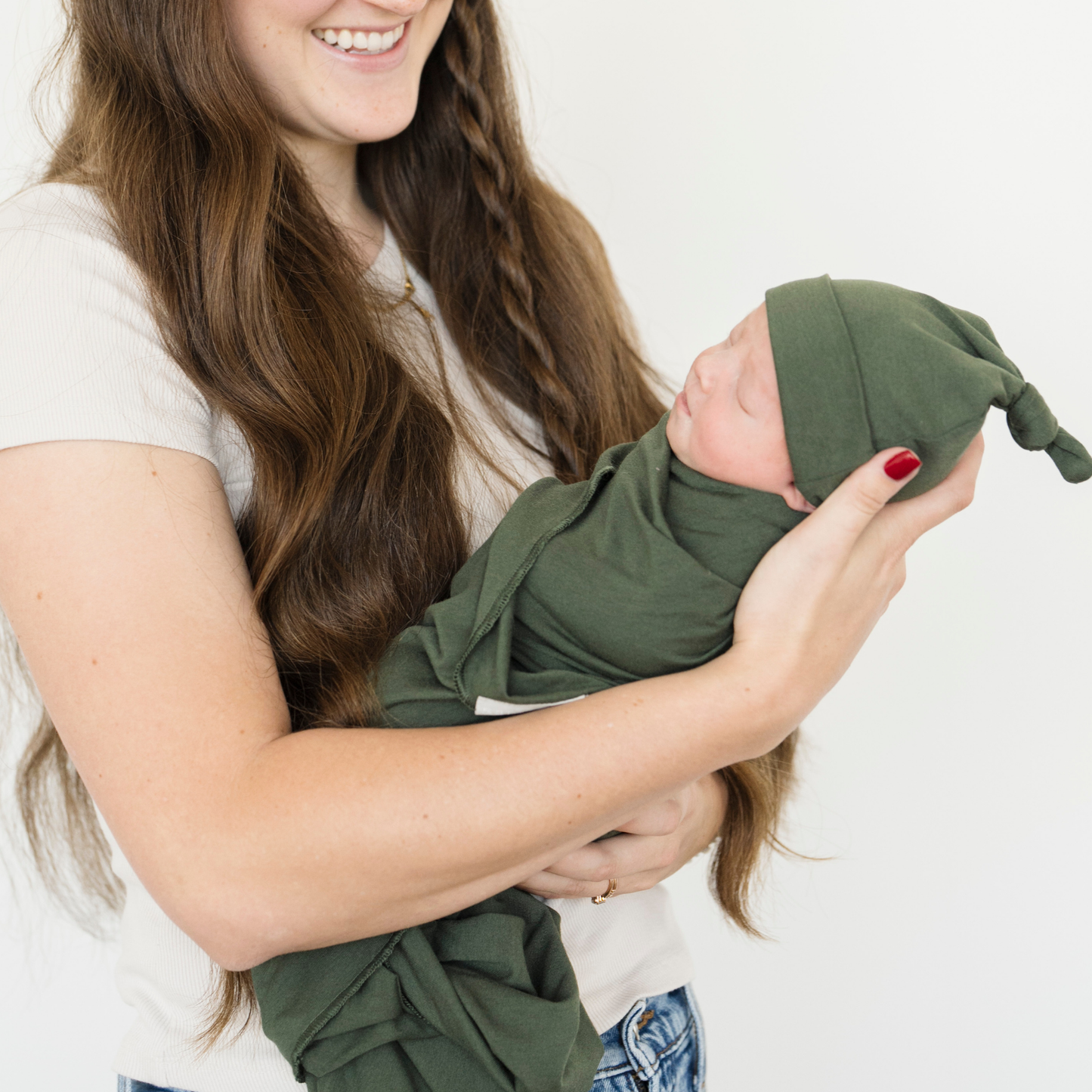 Mom holding baby girl swaddled in Juniper Stretchy Swaddle with a matching hat. 