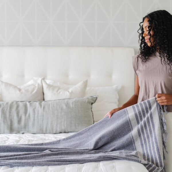 Woman with curly hair making a bed with a gray striped blanket - Saranoni