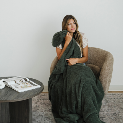 Woman sits in a chair snuggled with a juniper green bamboni extra large blanket.