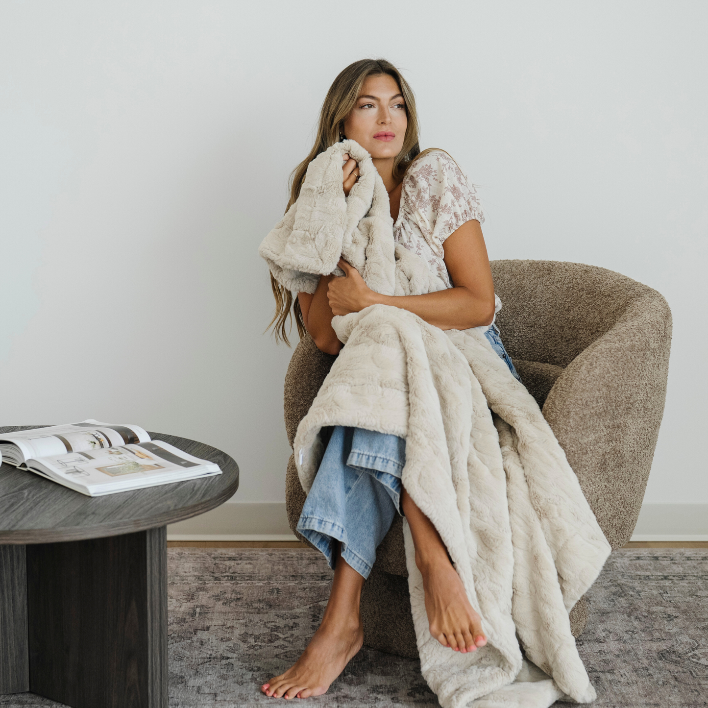 Woman sits in chair wrapped in a Buff cozy faux fur throw blanket. 