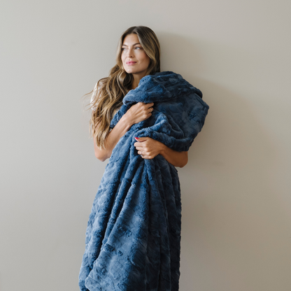 Woman stands snuggled with an indigo blue faux fur soft cozy blanket. 