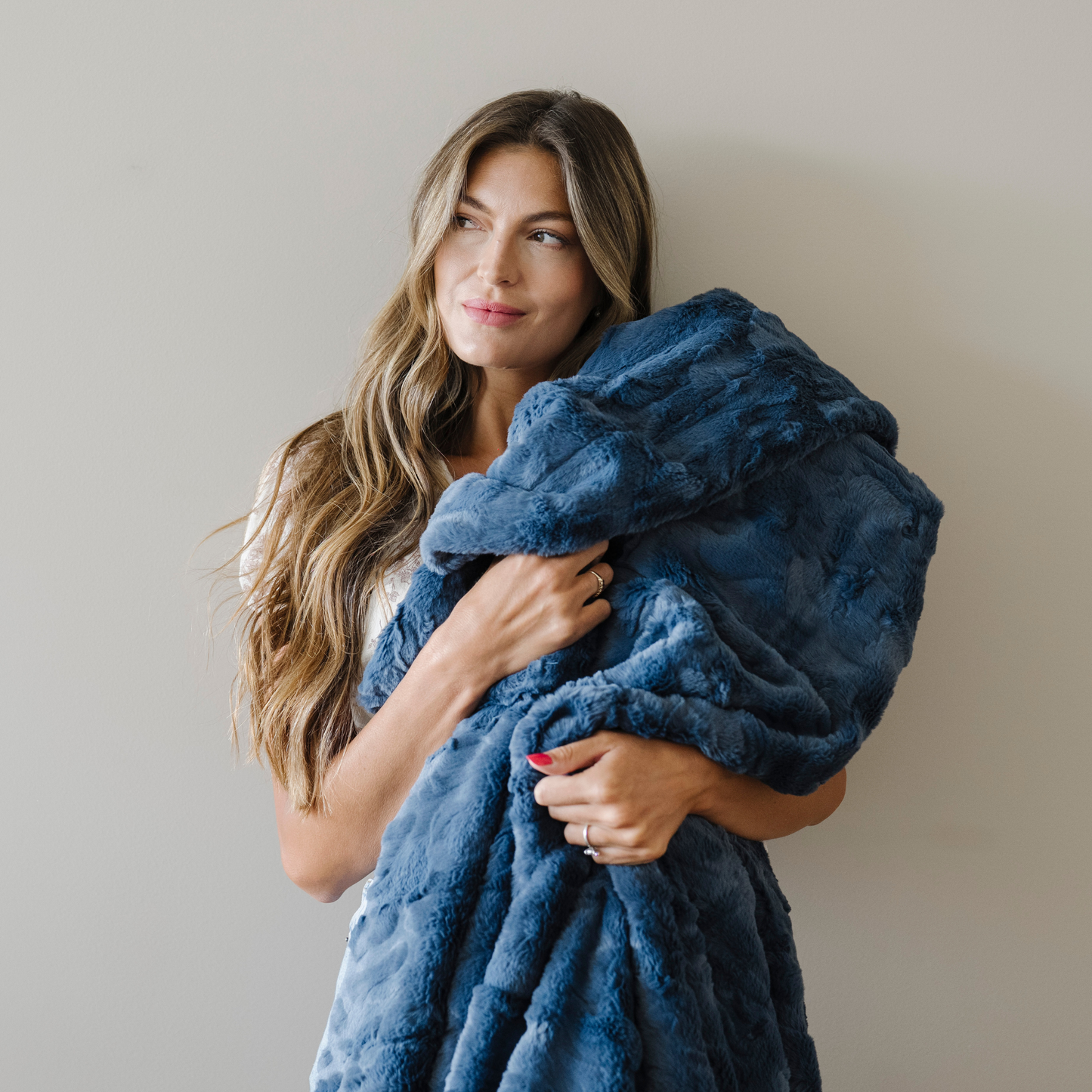Woman stands snuggled with a faux fur cozy throw blanket that is indigo blue. 