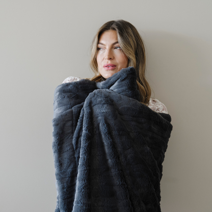 Woman is wrapped in a charcoal gray faux fur cozy throw blanket. 