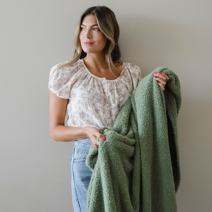 Woman stands holding an olive green cozy extra large bamboni blanket.