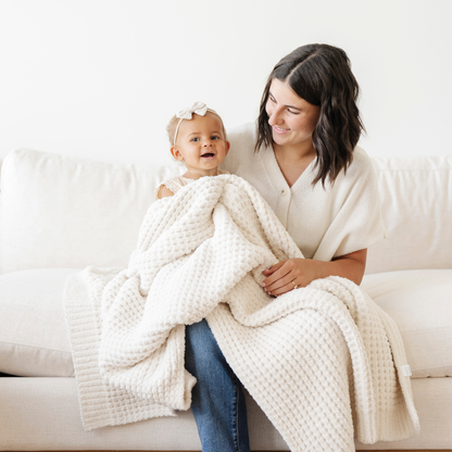Mom holds little girl snuggled with a cream waffle knit receiving baby blanket. 