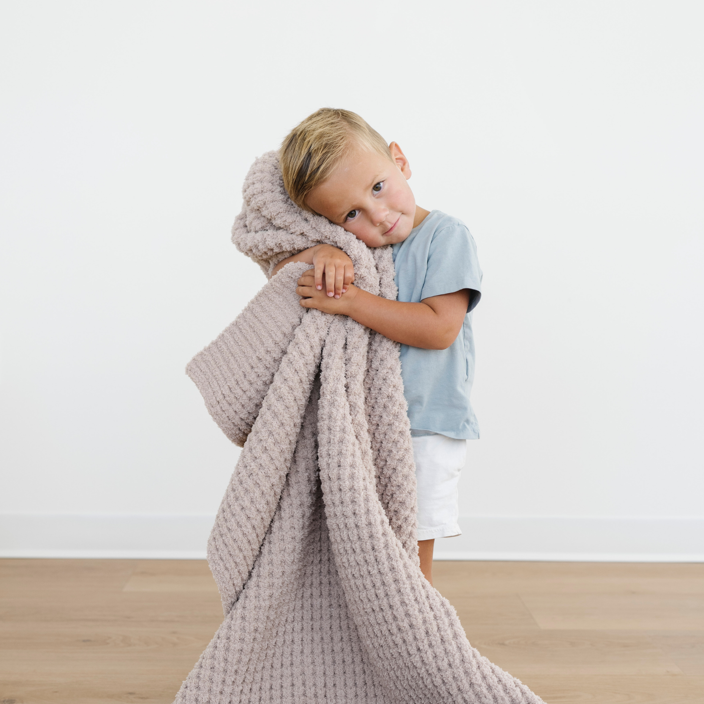 Little boy stands snuggling a tan waffle knit toddler blanket that is very soft and cozy. 
