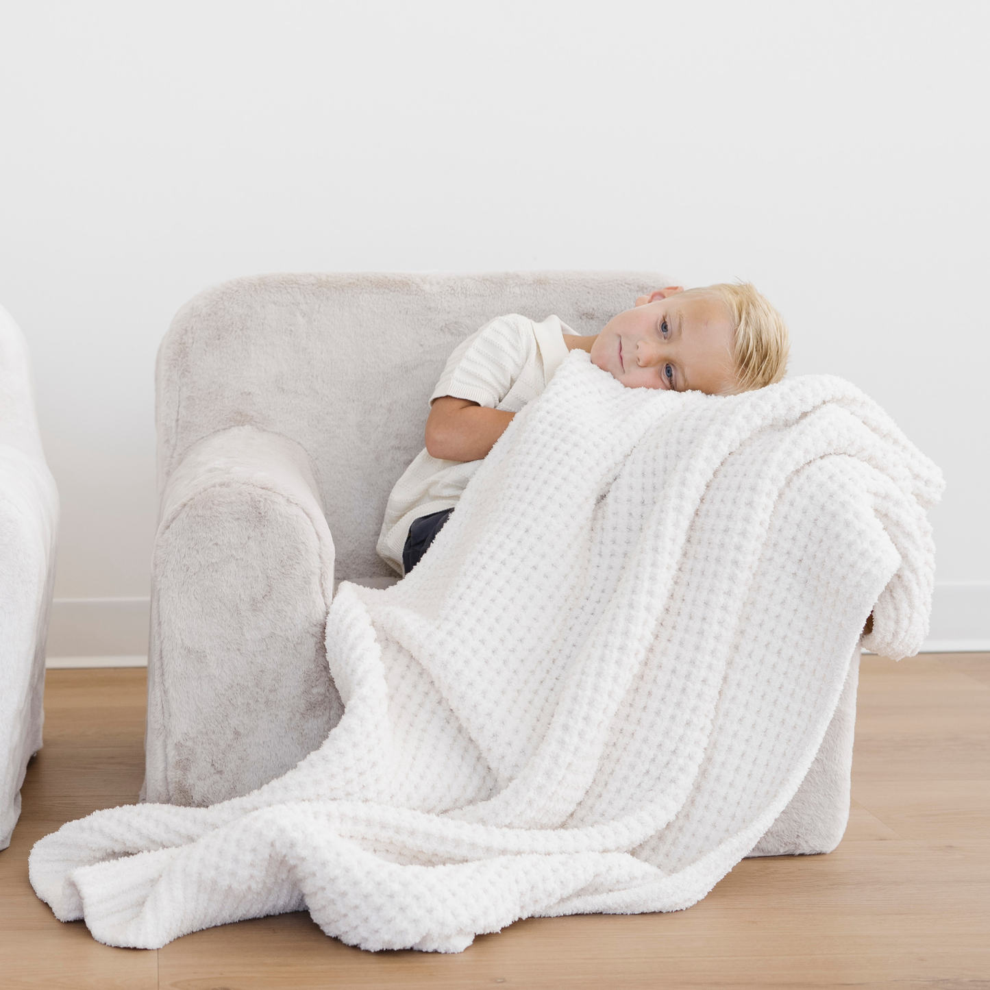Boy sits in a cozy soft chair snuggling a cream waffle knit toddler blanket. 