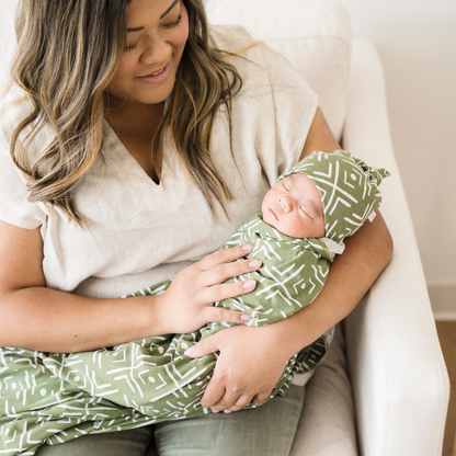 Mom holding baby boy swaddled in Olive Mudcloth Saranoni soft stretchy swaddle with a matching hat. 