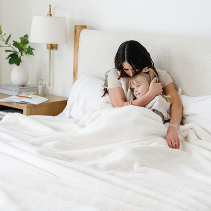 A mother and her daughter sit under a Saranoni Grand Faux Fur blanket, featuring an elegant white tone and luxuriously soft texture. This cozy, queen-sized blanket and faux fur blanket offers soothing weight, premium softness, timeless comfort, and is a soft blanket. Perfect as a big blanket and luxury blanket for snuggling or as a luxury bedroom accent, this Saranoni blanket elevates every cozy moment and is a cozy blanket.
