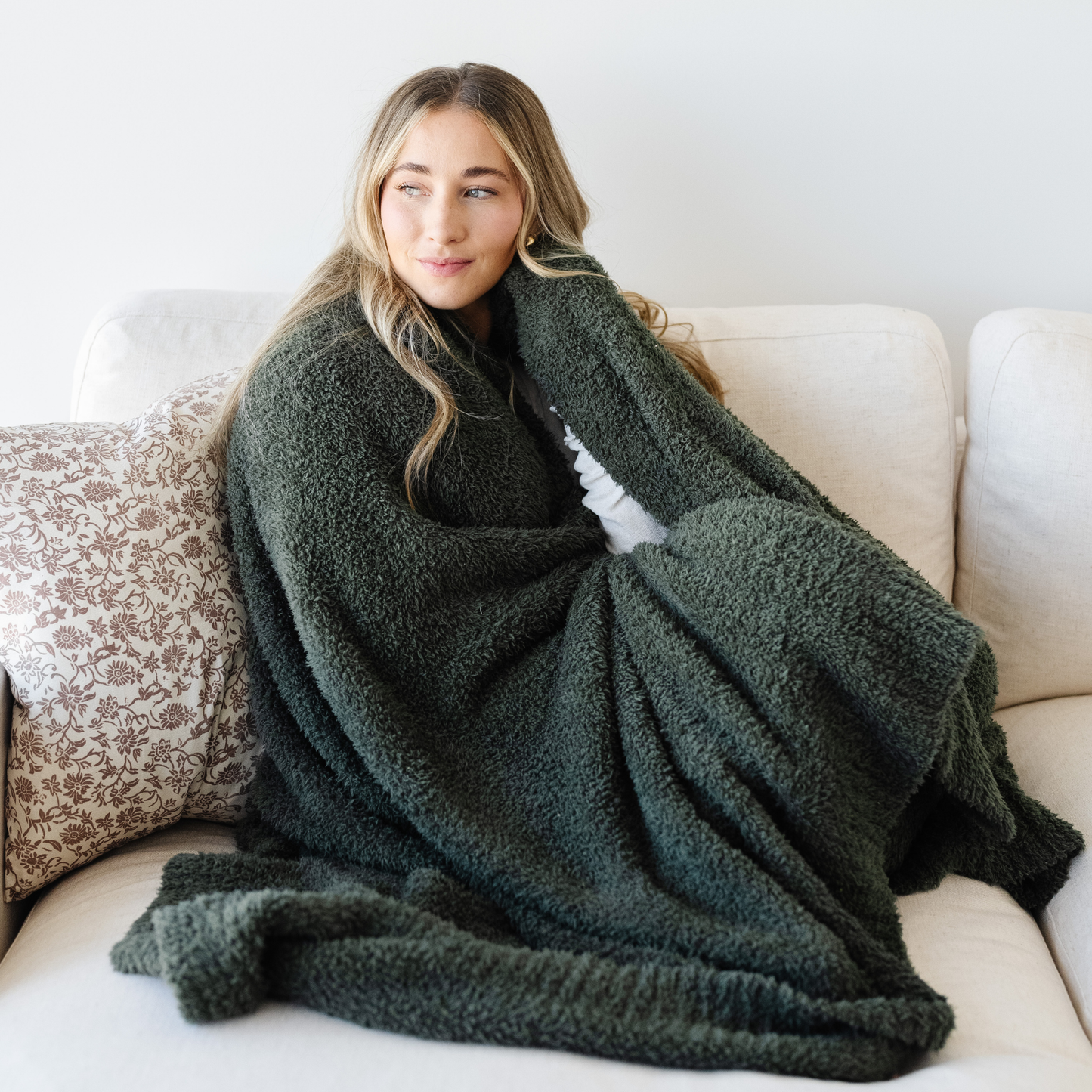 Woman sits on couch wrapped in a Juniper green bamboni blanket. 