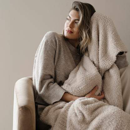 A woman is holding a light tan Bamboni® blanket and sitting in a chair. This soft blanket is a luxury blanket and a Saranoni blanket. This blanket's softness and breathability makes it a perfect comfort gift, home decor piece, or relaxing blanket. 