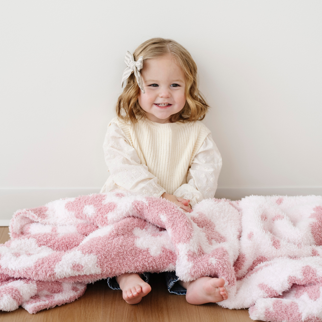 A smiling little girl is sitting with a receiving blanket that is a double-layer Bamboni® blanket. This soft blanket that is a Saranoni blanket is pink and light pink and has an adorable floral pattern. This is a great baby blanket or baby gift. This luxury blanket is also very breathable and stretchy.