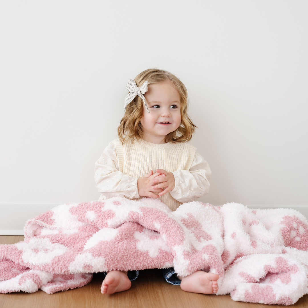 A little girl is sitting with a pink and light pink floral patterned double-layer Bamboni® blanket. This soft blanket is a luxury blanket and a Saranoni blanket. This also a great baby gift or baby blanket because it is a receiving blanket. 