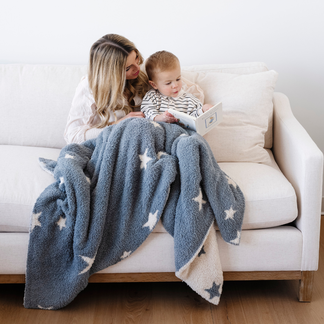 A mother and her son read a book while they are sitting on a couch with a Saranoni blanket. This soft blanket is a blue and cream double-layer Bamboni® blanket with an adorable star pattern. Since this kid's blanket is stretchy and durable, it makes it a great toddler gift.