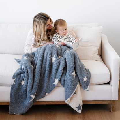 A mother and her son read a book while they are sitting on a couch with a Saranoni blanket. This soft blanket is a blue and cream double-layer Bamboni® blanket with an adorable star pattern. Since this kid's blanket is stretchy and durable, it makes it a great toddler gift.
