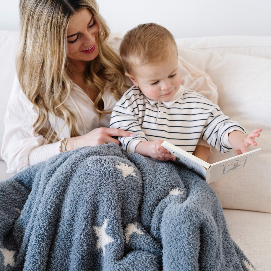 A mother and her little boy read a book while sitting on a couch. They have a double-layer Bamboni® blanket that is blue and cream with a star pattern on their laps. This soft blanket is a receiving blanket and a Saranoni blanket. The blanket's stretchiness and breathability make is a perfect baby gift and baby blanket. 