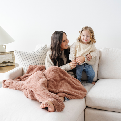 A mother and her little girl are on a couch with a pink Bamboni® blanket. This luxury extra large blanket is a Saranoni blanket and a cozy blanket. this blanket is great as a comfort gift, addition to your home decor, or for relaxing. Enjoy this soft blanket for years to come!