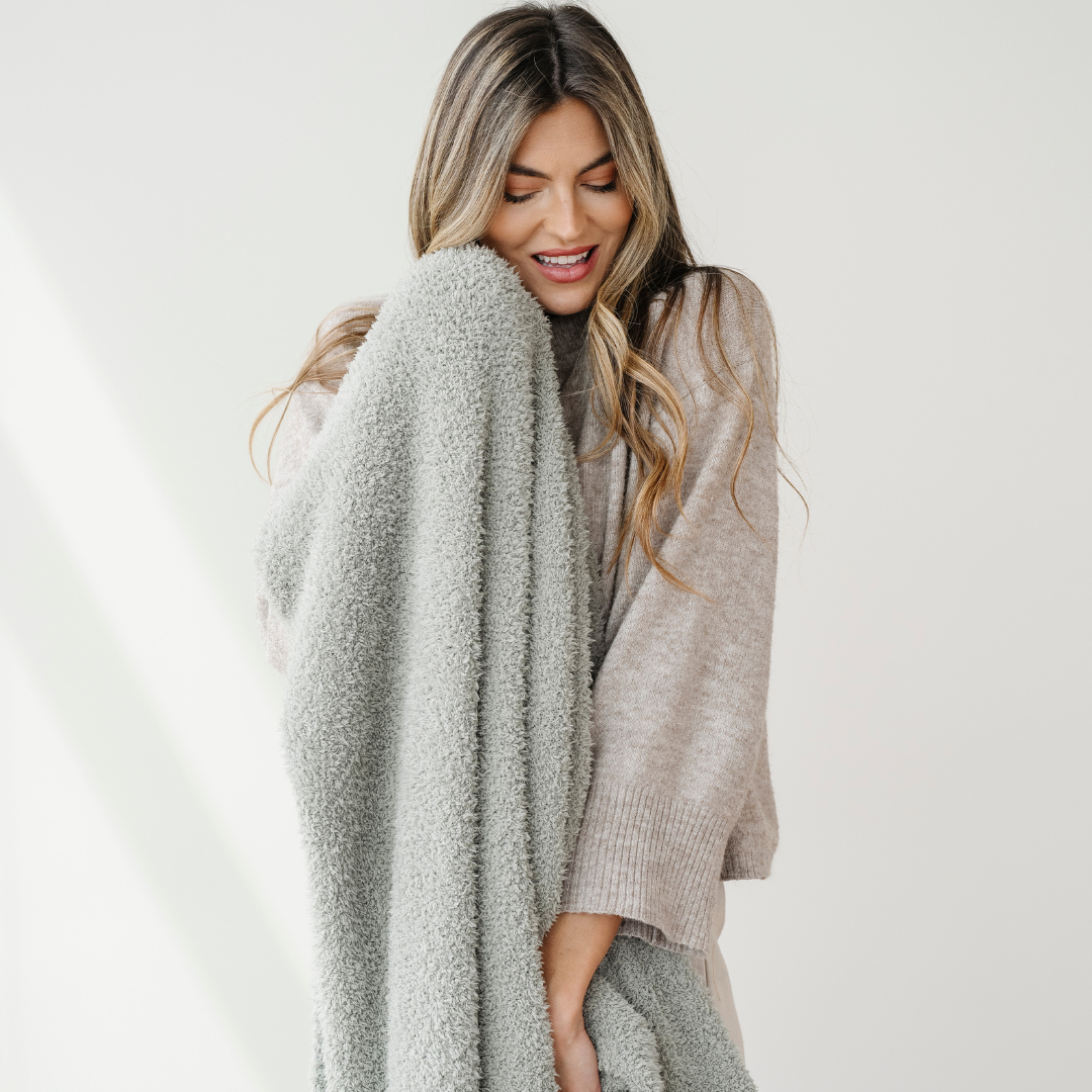 A woman is smiling and holding a sage colored Bamboni® blanket. This soft blanket is very stretchy and breathable. It's great for gifting, relaxing, or as a home decor item. This Saranoni blanket is a luxury blanket and an extra large blanket. 