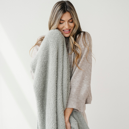 A woman is smiling and holding a sage colored Bamboni® blanket. This soft blanket is very stretchy and breathable. It's great for gifting, relaxing, or as a home decor item. This Saranoni blanket is a luxury blanket and an extra large blanket. 