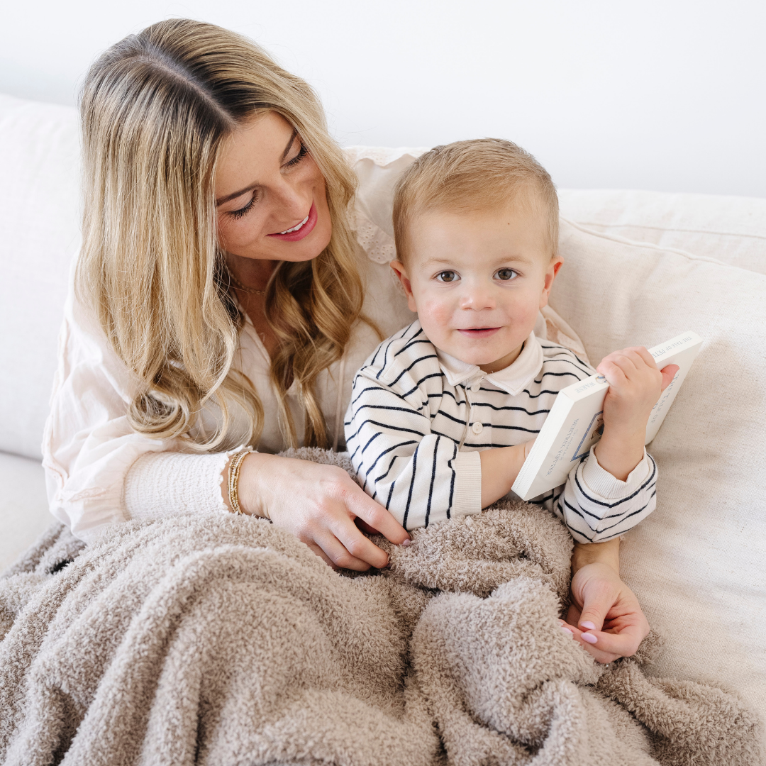 A mother and her son are sitting on a couch and there is a taupe Bamboni® toddler blanket that is a Saranoni blanket. This cozy blanket is a great gift for toddlers or a addition to a bedroom. This soft blanket is also very stretchy and breathable. 