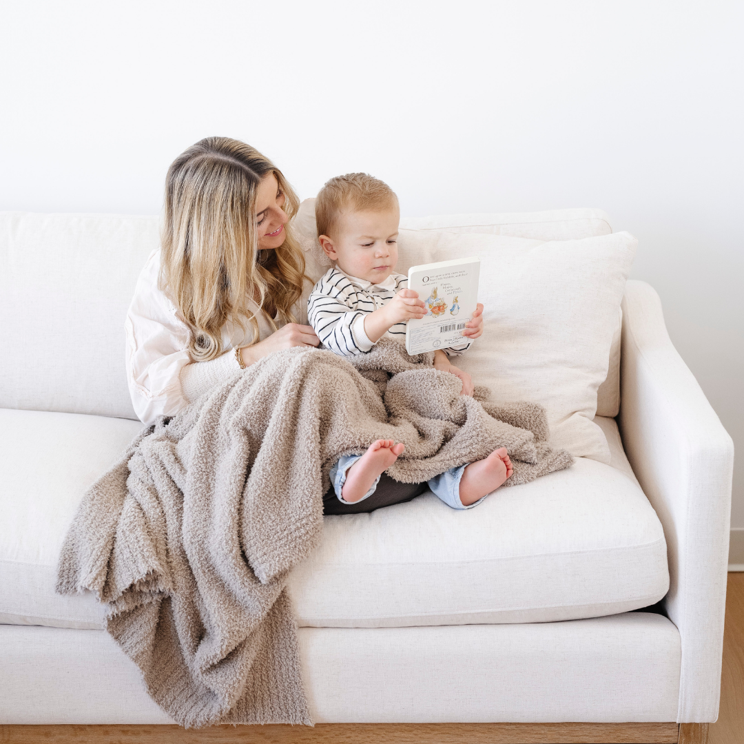 A mother and her son sit on a couch. They have a taupe Bamboni® toddler blanket on their laps. This cozy blanket is a Saranoni blanket and is very stretchy and breathable. This kid's blanket is a great gift for toddlers or addition to a bedroom. 