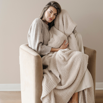 A woman is sitting in a chair and holding a light tan blanket that is a Bamboni® blanket. This soft blanket is very stretchy and breathable. This Saranoni blanket is also great for gifting, relaxing, or as a home decor staple. 