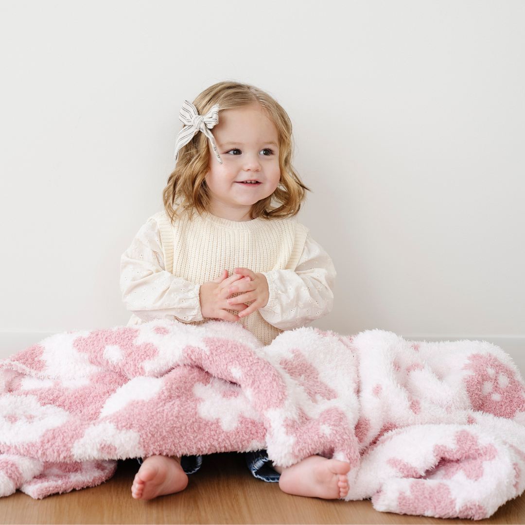 A little girl is sitting with a toddler blanket that is pink and light pink with an adorable floral pattern. This double-layer Bamboni® blanket is a Saranoni blanket and is so soft. This is a great gift for toddlers because it is stretchy and breathable. 
