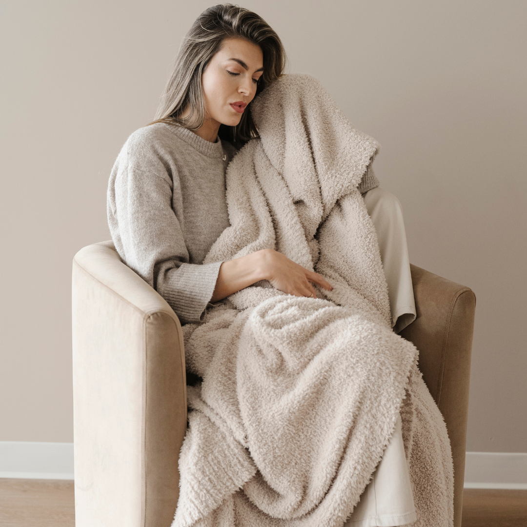 A woman is sitting with a light tan Bamboni® blanket that is an extra-large blanket. This soft blanket is a Saranoni blanket that is stretchy and breathable. Use this cozy blanket for relaxing, gifting, or as a home decor piece. Enjoy for years to come. 